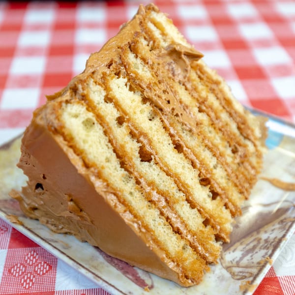 A slice of layered cake with light brown frosting on a patterned plate over a red and white checkered tablecloth.