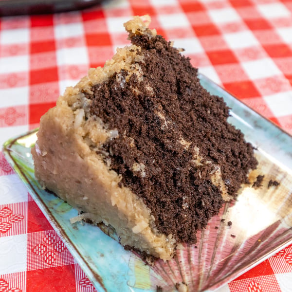 A slice of chocolate cake with coconut frosting on a decorative plate over a red and white checkered tablecloth.