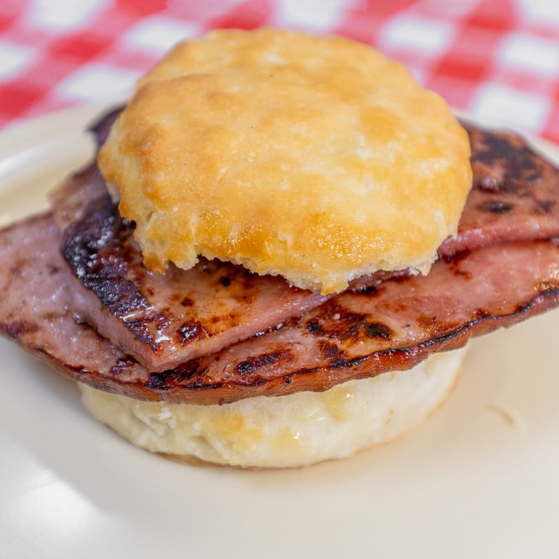 Close-up of a golden-brown biscuit sandwich with grilled meat on a white plate and red checkered background.