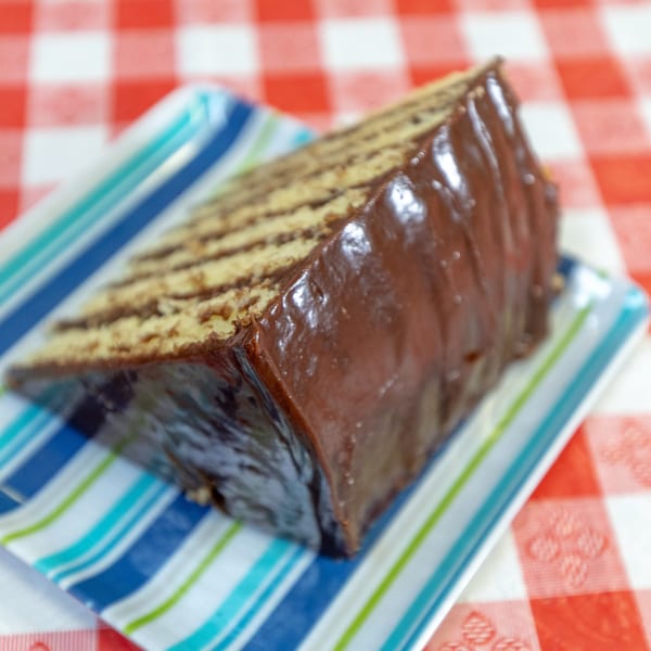 A slice of chocolate cake with many layers, served on a striped plate over a red and white checkered tablecloth.