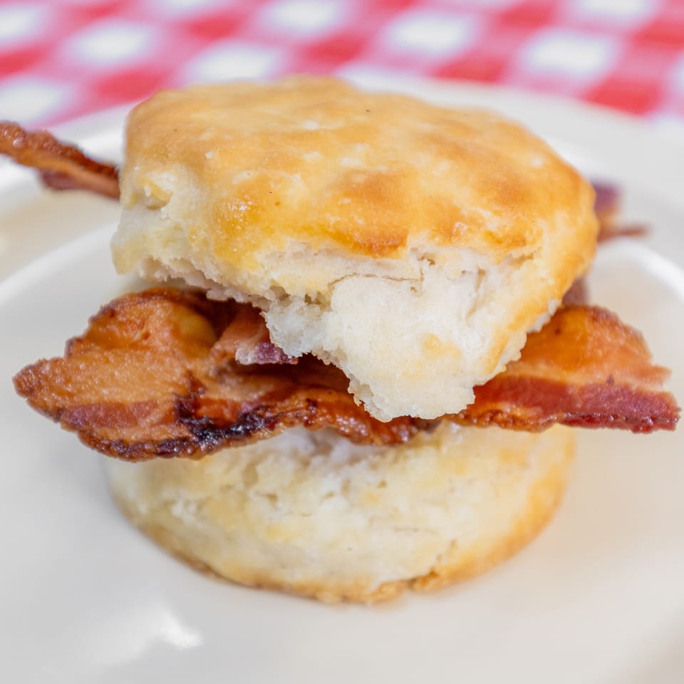 A golden-brown biscuit sandwich with crispy bacon on a white plate with a red and white checkered background.