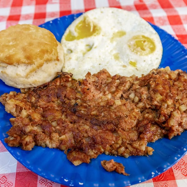 A blue plate holds a golden biscuit, two fried eggs, and a large serving of corned beef hash.