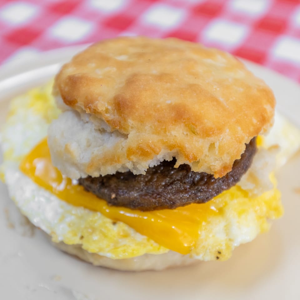 Close-up of a breakfast biscuit sandwich with sausage, egg, and cheese on a white plate.