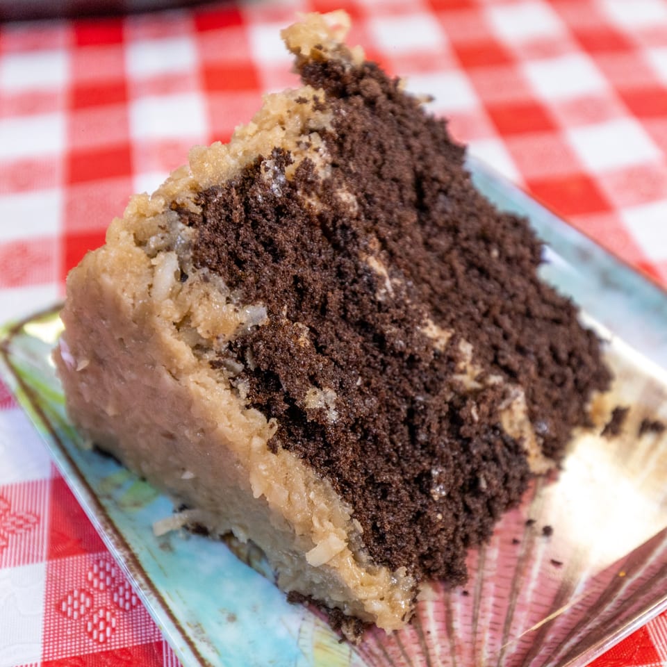 A slice of chocolate cake with coconut pecan frosting on a decorative plate over a red and white checkered tablecloth.