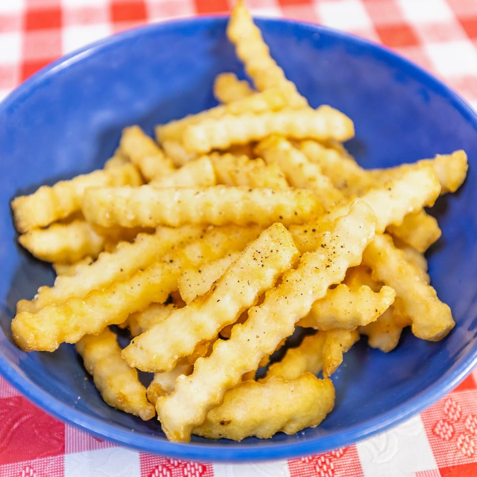 A blue bowl filled with golden, crinkle-cut french fries seasoned with pepper on a red and white checkered tablecloth.