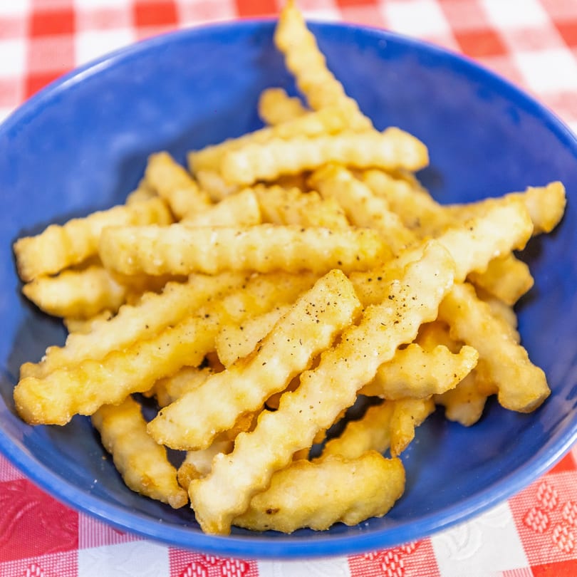 A blue bowl filled with golden, crinkle-cut french fries seasoned with pepper on a red and white checkered tablecloth.