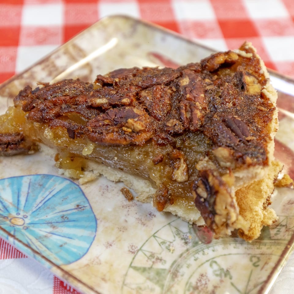 A slice of pecan pie with a golden-brown crust and gooey filling on a decorative plate.